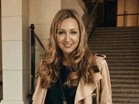 Smiling person with long hair wearing a coat, standing indoors near stairs.