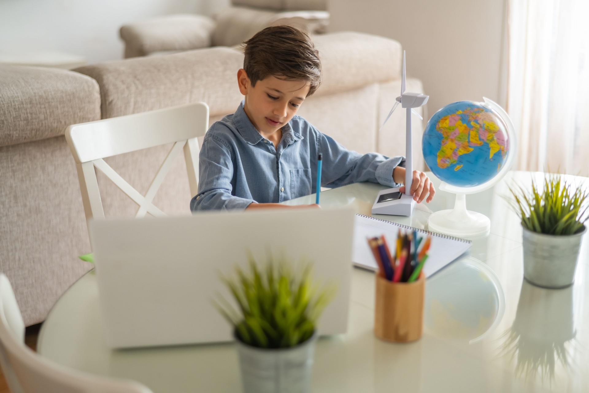 Elementary age schoolboy is learning about renewable energy at home, using a small wind turbine model, a globe, and a laptop for his stem education project