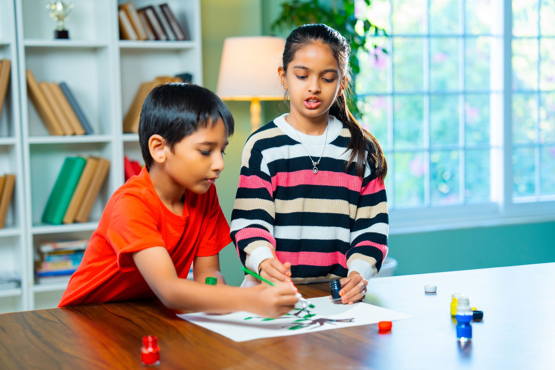 Indian siblings exploring creativity by painting on a drawing paper at home, using brushes and colours