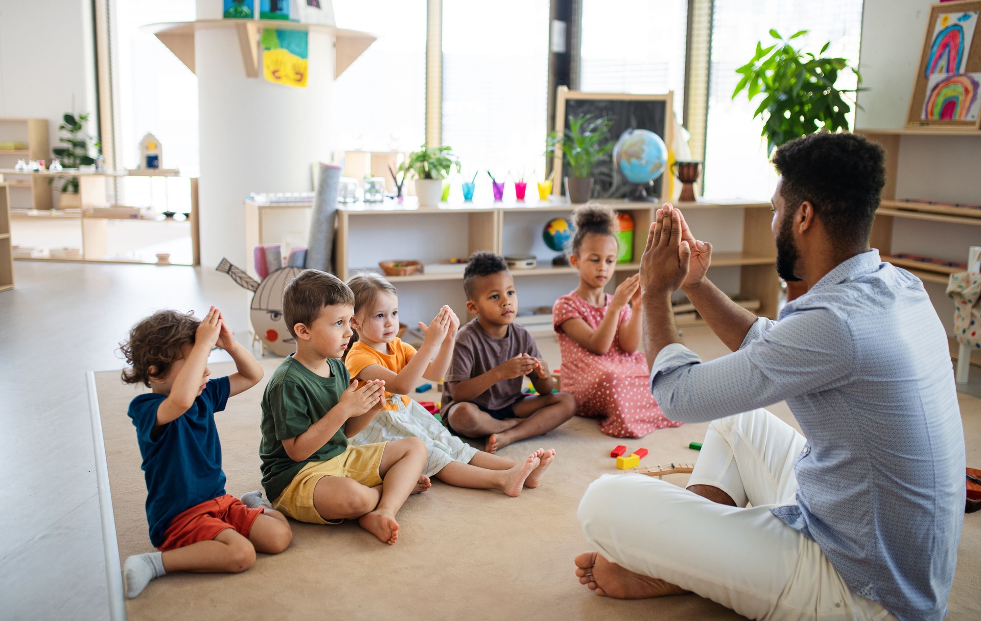 Group of small nursery school children with man teacher sitting on floor indoors in classroom, playing.